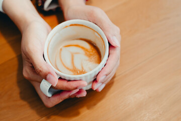 Hands Holding a White Mug with Coffee and Art Design on the Surface, Warm Atmosphere in a Cozy Coffee Shop Setting