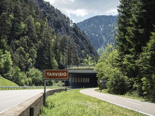 A scenic bicycle path near Tarvisio, Italy, winding through lush alpine forests and mountains. A road sign marks the town's entrance, with a tunnel in the background.
