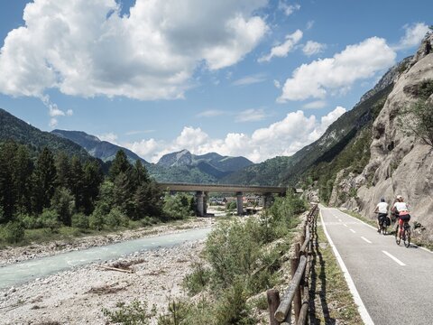 A paved cycling path along the Alpe Adria Radweg in Italy, surrounded by lush green mountains and a scenic river. A bridge spans the valley, while a cyclist enjoys the peaceful landscape.