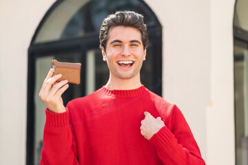 Young handsome man holding a wallet at outdoors with surprise facial expression