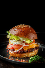 A delicious, layered hamburger featuring a sesame seed bun, grilled patty, fresh lettuce, tomato, pickles, onions, and crunchy tortilla chips, placed on a black plate.