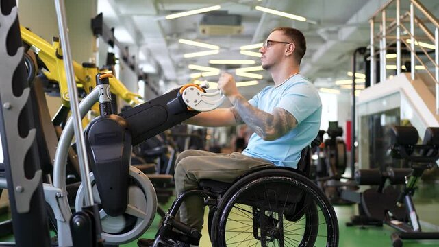 Disabled sportive man in wheelchair works on upper body muscles in gym. Injury from accident, strong spirit, dedication to sports and health, using arm cycle trainer for rehabilitation. Profile view