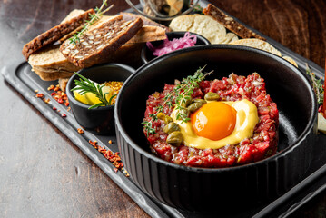 A hearty dish of steak tartare topped with a raw egg yolk, surrounded by garnishes, served with crispy French fries, toasted bread, and pickled onions on a wooden table.