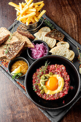 A hearty dish of steak tartare topped with a raw egg yolk, surrounded by garnishes, served with crispy French fries, toasted bread, and pickled onions on a wooden table.