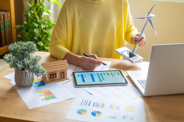 Businesswoman analyzing charts on a digital tablet, evaluating data for renewable energy investments alongside miniature wind turbine and solar panel models