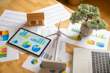 Desk displaying a miniature house and wind turbine model alongside charts, graphs, and a digital tablet, highlighting renewable energy investment and sustainable development