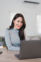 Asian businesswoman in white shirt sits at desk, smiling while using smartphone, with laptop and coffee mug nearby.