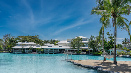 Swimming pool in a tropical resort. The hammock is suspended from the trunks of palm trees in the recreation area. A canopy by the aquamarine water. White cottages among the green vegetation. Blue sky