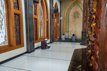 A serene interior of a mosque with intricate Islamic architecture, where three men are engaged in prayer and reading the Quran in a peaceful setting.