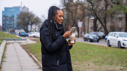 Fototapeta premium Young african american woman in the morning enjoying daily walk in the city, walking on the street texting on smartphone, holding coffee cup.