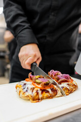 A chef in a black shirt is slicing a freshly baked pizza on a wooden board, showcasing vibrant toppings like onions, peppers, and melted cheese.