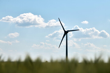 Single wind turbine silhouetted against bright blue sky with scattered fluffy clouds and blurred...