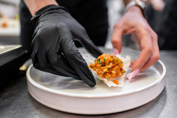 A chef's hand in a black glove carefully places a colorful mix of diced ingredients onto a delicate rice paper wrap, showcasing culinary artistry in a well-lit kitchen.
