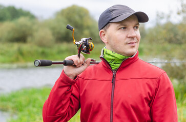 Fisherman in red sweatshirt and cap on the river bank with spinning rod in his hands.