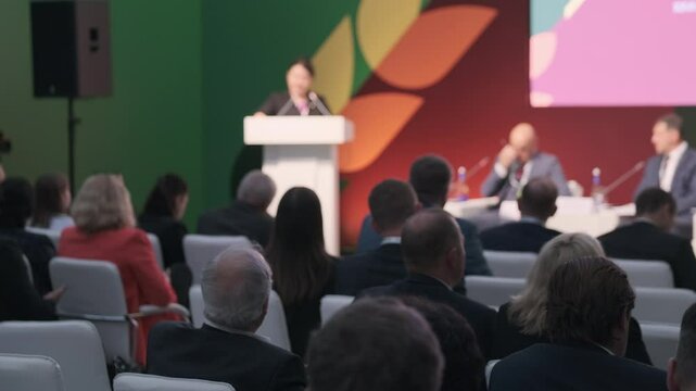 Meeting of individuals in a conference with speakers, audience, white chairs, and bright background.