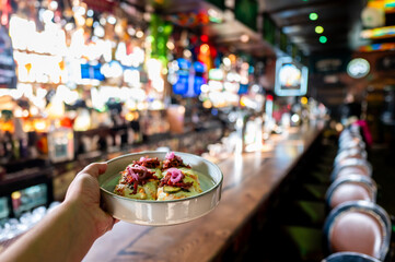 A hand holding a plate of delicious food in a lively bar, with a vibrant and illuminated background showcasing drinks and a bustling atmosphere.