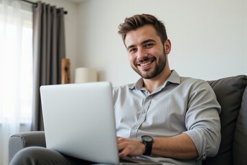 A Smiling Young Caucasian Man in Casual Attire Working on a Laptop While Sitting Comfortably on a Gray Couch in a Bright, Modern Living Room