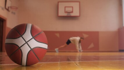 Basketball ball lies on wooden floor of gym with player stretching in background - Powered by Adobe