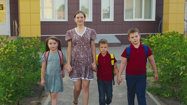 Mother leading children from school, female mother holding children hand, happy family, young nanny with children, schoolchildren with backpacks walking with aunt by the hand, little boys girl walking