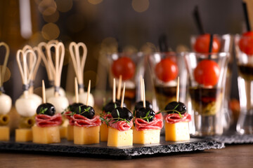 Many different tasty canapes on wooden table against blurred lights, closeup