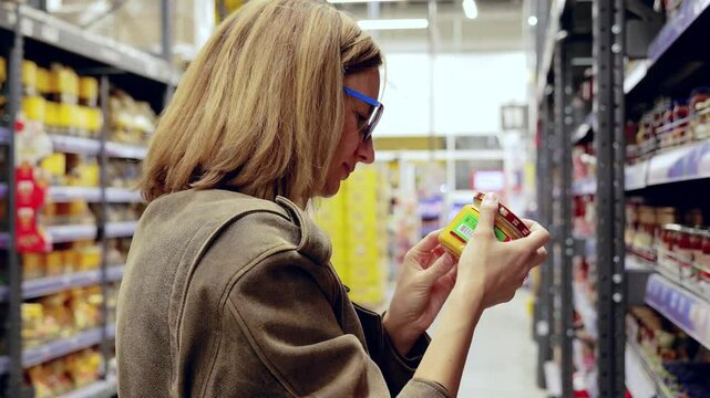 Woman in glasses and a brown jacket is shopping in a supermarket, carefully reading the label of a jar in the baby food aisle