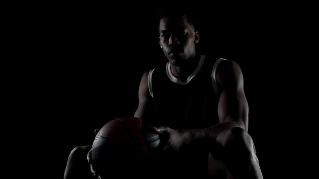 Basketball player sitting on a bench. Looking at camera, Concentrated african american sports man against black background.