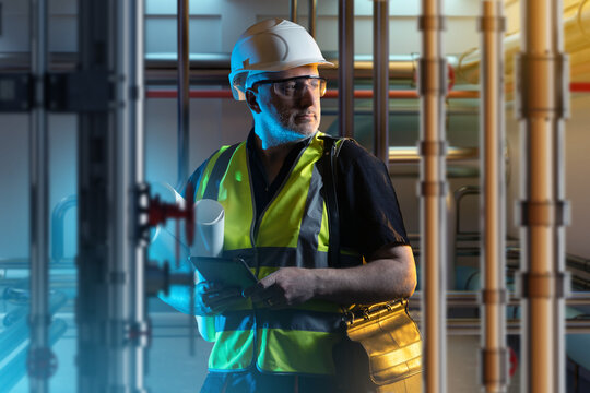 Boiler room engineer. Man among pipes. Human in engineers clothes is standing indoor. Guy in room with utilities. Boiler room design concept. Worker with tablet among boiler equipment. Art focus.