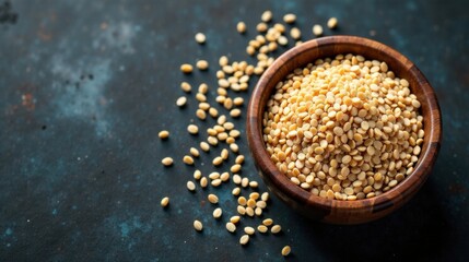 A wooden bowl overflowing with pale yellow split peas, scattered on a dark textured surface.