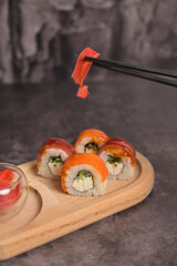Set of sushi roll with rice and fish on wooden serving board. Chopsticks holding ginger. Dark black texture background with copy space. Japan menu. Asian food