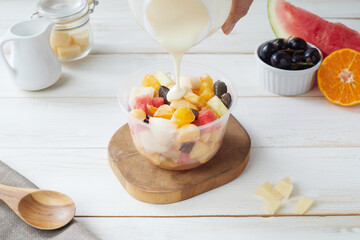 Fruit Salad Being Prepared in a Plastic Bowl With Fresh Ingredients, salad dressing and grated cheese on a Wooden Table