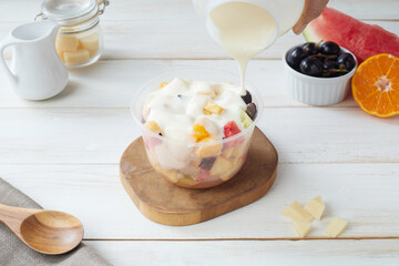 Fresh Fruit Salad in a Plastic Bowl Topped With Creamy Dressing on a Wooden Board in a Bright Kitchen Setting