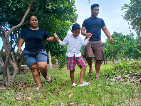 A family of three, including a woman, a man, and a boy, is walking hand in hand in a garden with lush green trees. The boy is in the center, wearing a white shirt and red checkered shorts.