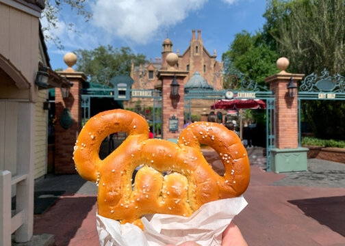 A Mouse Ear Shaped Pretzel Held In Front Of A Theme Park Attraction