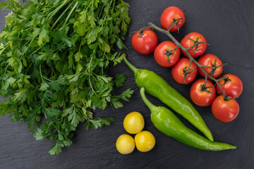 Fresh parsley, cherry tomatoes on the vine, green chili peppers, and yellow cherry plums on a dark slate background. The ingredients are arranged in a balanced composition
