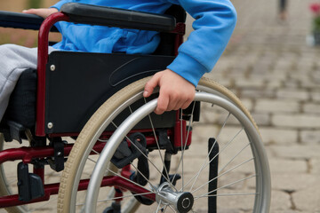 Fototapeta premium A close up of a child’s hand gripping the wheel of a wheelchair while wearing a blue hoodie, moving on a cobblestone path outdoors.