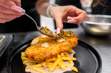 A close-up of a hand with well-manicured nails adding toppings to a crispy fried chicken piece on a bed of fries. The dish is plated on a black plate, showcasing a delicious meal.