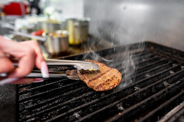 A close-up view of a hand using tongs to flip a sizzling hamburger patty on a smoky grill, showcasing the texture and juiciness of the meat.
