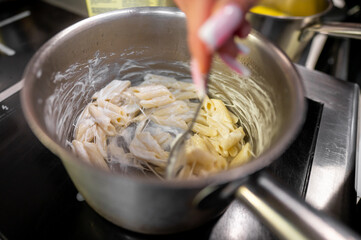 A close-up of a hand stirring pasta in a saucepan, coated with creamy sauce, on a stove. The scene depicts a delicious cooking moment in a kitchen setting.