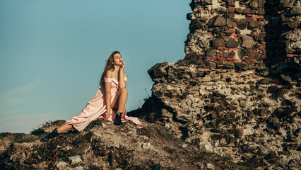 A woman in a long pink dress sits on a rocky surface next to a destroyed stone wall. She looks thoughtfully into the distance, placing her hand on her chin. The background is clear blue sky.