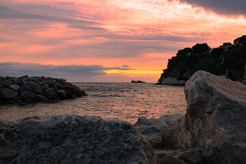 Seascape, view of the sea surface with a ship sailing in the distance, large stones in the foreground, and in the background - the sky, painted with the colours of a beautiful sunset. 