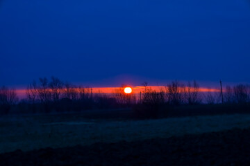 The photo depicts a sunset over the horizon. The sun, bright orange, is in the center of the frame,...