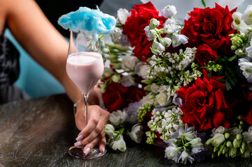 A elegantly styled drink topped with blue cotton candy is held by a hand, surrounded by a vibrant bouquet of red and white flowers on a dark wooden table.