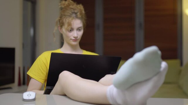 Girl working on a laptop in her lap. Casual clothing with feet up on the table in a living room at home. Portrait of a student taking online course. Home education. Focus on different color socks.