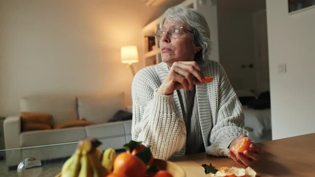 A happy senior woman enjoys a healthy snack of fresh mandarin or oranges in her cozy home, promoting healthy aging and wellbeing.