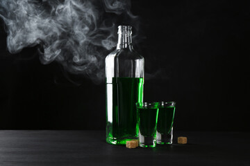 Absinthe in shot glasses, bottle and brown sugar on table against black background with smoke