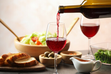 Pouring red wine into glass at grey table with tasty food, closeup