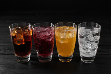 Refreshing soda water of different flavors with ice cubes in glasses on black wooden table