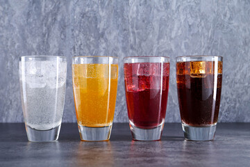 Sweet soda water of different flavors with ice cubes in glasses on grey table