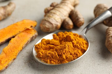 Aromatic turmeric powder in spoon and fresh roots on light grey table, closeup