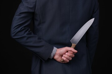 Businessman holding knife behind his back on black background, closeup